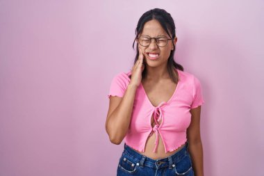 Hispanic young woman standing over pink background wearing glasses touching mouth with hand with painful expression because of toothache or dental illness on teeth. dentist 