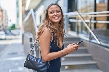 Genç, güzel, İspanyol bir kadın sokakta akıllı telefon kullanarak gülümsüyor.