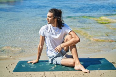 Young hispanic man doing yoga exercise sitting on sand at beach