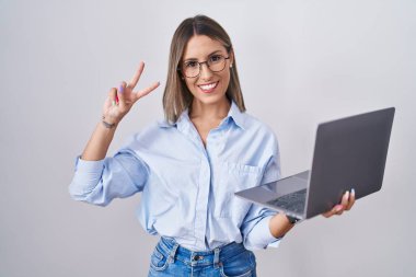 Young woman working using computer laptop smiling looking to the camera showing fingers doing victory sign. number two. 