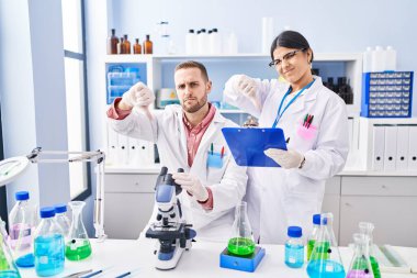 İki genç bilim adamı, kızgın yüzüyle laboratuvarda çalışıyor. Negatif işaretler, baş parmakları aşağıda, reddedilme kavramı. 