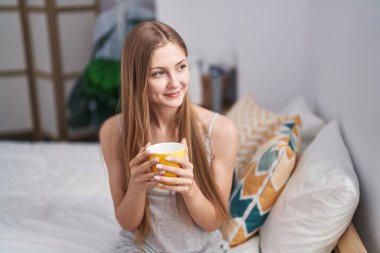 Young caucasian woman drinking cup of coffee sitting on bed at bedroom
