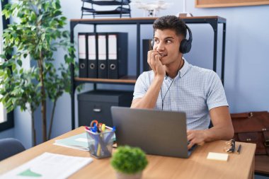 Young hispanic man working at the office wearing headphones looking stressed and nervous with hands on mouth biting nails. anxiety problem. 