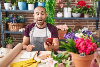 Hispanic man with beard working at florist shop with smartphone scared and amazed with open mouth for surprise, disbelief face 