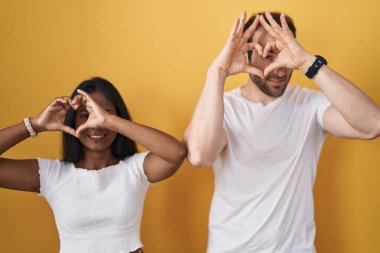 Interracial couple standing over yellow background doing heart shape with hand and fingers smiling looking through sign 