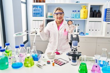 Young hispanic woman working at scientist laboratory doing make up surprised with an idea or question pointing finger with happy face, number one 
