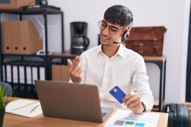Young hispanic man working using computer laptop holding credit card beckoning come here gesture with hand inviting welcoming happy and smiling 