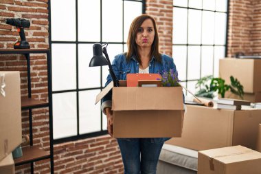 Middle age woman moving to a new home holding cardboard box puffing cheeks with funny face. mouth inflated with air, catching air. 
