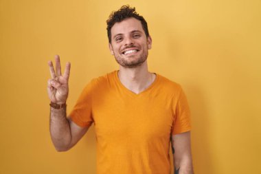 Young hispanic man standing over yellow background showing and pointing up with fingers number three while smiling confident and happy. 