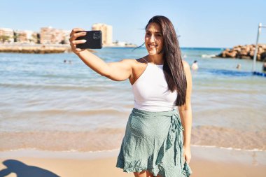 Genç İspanyol kadın gülümsüyor. Deniz kenarındaki akıllı telefonun yanında özçekim yapıyor.