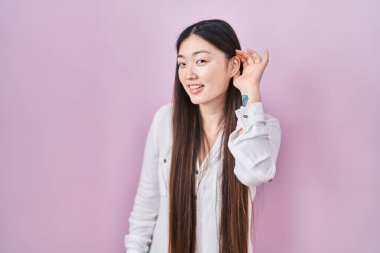 Chinese young woman standing over pink background smiling with hand over ear listening an hearing to rumor or gossip. deafness concept. 