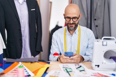 Genç kel adam terzide akıllı telefon kullanarak gülümsüyor.
