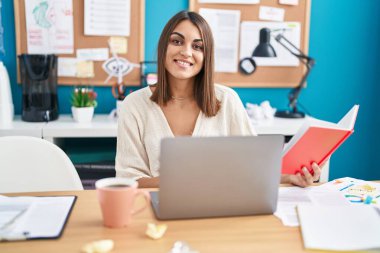 Young beautiful hispanic woman business worker using laptop reading notebook at office