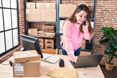 Young beautiful hispanic woman ecommerce business worker using laptop talking on smartphone at office