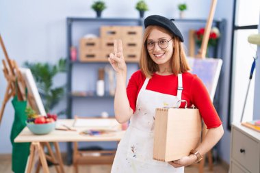 Young redhead woman at art studio holding art case smiling with happy face winking at the camera doing victory sign with fingers. number two. 