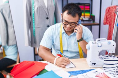 Young chinese man tailor talking on smartphone writing on notebook at tailor shop