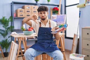 Arab man with beard painter sitting at art studio holding palette with angry face, negative sign showing dislike with thumbs down, rejection concept 