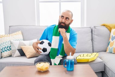 Young hispanic man with beard and tattoos football hooligan holding ball supporting team serious face thinking about question with hand on chin, thoughtful about confusing idea 