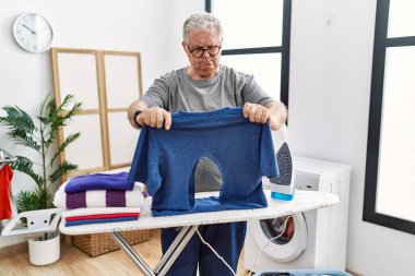 Senior caucasian man ironing holding burned iron shirt at laundry room depressed and worry for distress, crying angry and afraid. sad expression. 