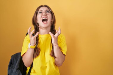 Sarı arka planda öğrenci çantası takan beyaz bir kadın çılgın ve sinirli bir şekilde bağırıyor ve agresif bir ifade ve kolla bağırıyor. hüsran kavramı. 