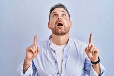 Mavi arka planda duran orta yaşlı beyaz adam şaşırmış ve şaşırmış bir şekilde yukarı baktı ve parmaklarıyla işaret ederek kollarını kaldırdı.. 