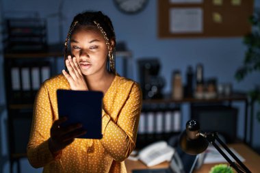 African american woman with braids working at the office at night with tablet hand on mouth telling secret rumor, whispering malicious talk conversation 