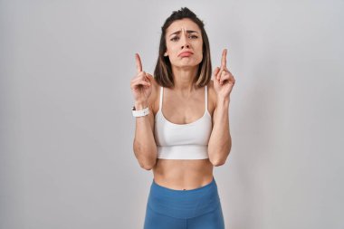 Hispanic woman wearing sportswear over isolated background pointing up looking sad and upset, indicating direction with fingers, unhappy and depressed. 