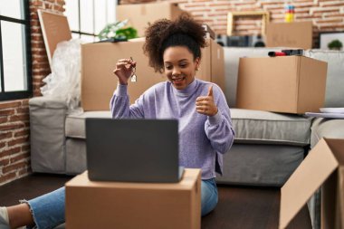 Young african american woman showing keys of new home on video call smiling happy and positive, thumb up doing excellent and approval sign 