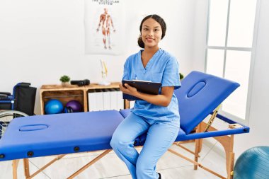 Fizyoterapist üniforması giyen genç Latin bir kadın klinikteki panoya yazı yazıyor.