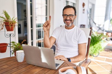 Middle age man using computer laptop at home showing and pointing up with finger number one while smiling confident and happy. 