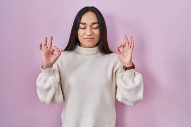 Young south asian woman standing over pink background relaxed and smiling with eyes closed doing meditation gesture with fingers. yoga concept. 