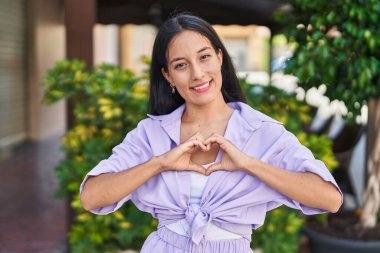 Genç, güzel, İspanyol bir kadın gülümsüyor. Kendine güveniyor, elleriyle kalp hareketi yapıyor.