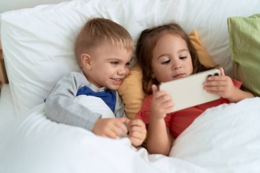 Adorable girl and boy watching video on smartphone lying on bed at bedroom