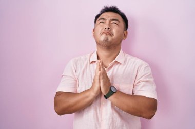 Chinese young man standing over pink background begging and praying with hands together with hope expression on face very emotional and worried. begging. 