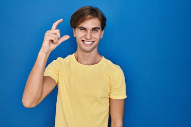 Young man standing over blue background smiling and confident gesturing with hand doing small size sign with fingers looking and the camera. measure concept. 