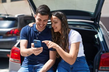Young hispanic couple sitting on car trunk using smartphone at street