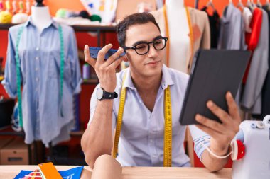 Young hispanic man tailor listening voice message by smartphone using touchpad at atelier