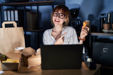 Young beautiful woman working using computer laptop and eating delivery food with a big smile on face, pointing with hand finger to the side looking at the camera. 