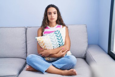Young hispanic woman sitting on the sofa at home relaxed with serious expression on face. simple and natural looking at the camera. 