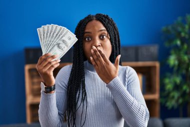 African american woman holding dollars banknotes covering mouth with hand, shocked and afraid for mistake. surprised expression 