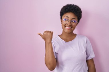 Young african american woman standing over pink background smiling with happy face looking and pointing to the side with thumb up. 