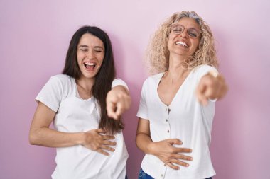 Anne ve kızı pembe arka planda yan yana durmuş sana gülüyorlar, parmaklarını kameraya doğru uzatıyorlar, yüz ifadesi utanç verici. 