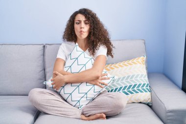 Hispanic woman with curly hair sitting on the sofa at home making fish face with mouth and squinting eyes, crazy and comical. 