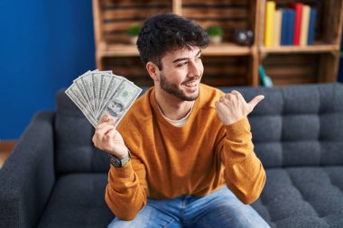 Hispanic man with beard holding 100 dollars banknotes pointing thumb up to the side smiling happy with open mouth 