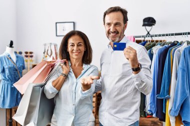 Hispanic middle age couple holding shopping bags and credit card smiling cheerful offering palm hand giving assistance and acceptance. 