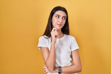 Young beautiful woman standing over yellow background with hand on chin thinking about question, pensive expression. smiling with thoughtful face. doubt concept. 