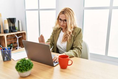 Beautiful blonde woman working at the office with laptop doing stop sing with palm of the hand. warning expression with negative and serious gesture on the face. 