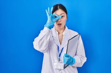 Chinese young woman working at scientist laboratory doing ok gesture shocked with surprised face, eye looking through fingers. unbelieving expression. 