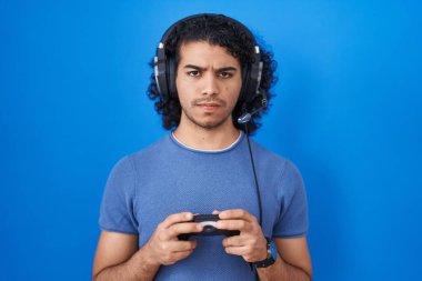 Hispanic man with curly hair playing video game holding controller skeptic and nervous, frowning upset because of problem. negative person. 