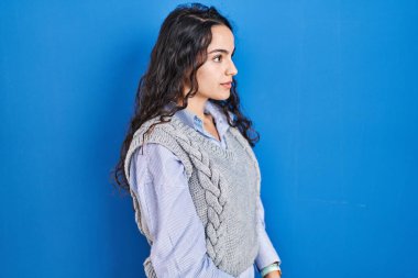 Genç esmer kadın mavi arka planda duruyor, yan tarafa bakıyor, rahat poz veriyor doğal bir yüzü ve kendine güvenen bir gülümsemesi var.. 